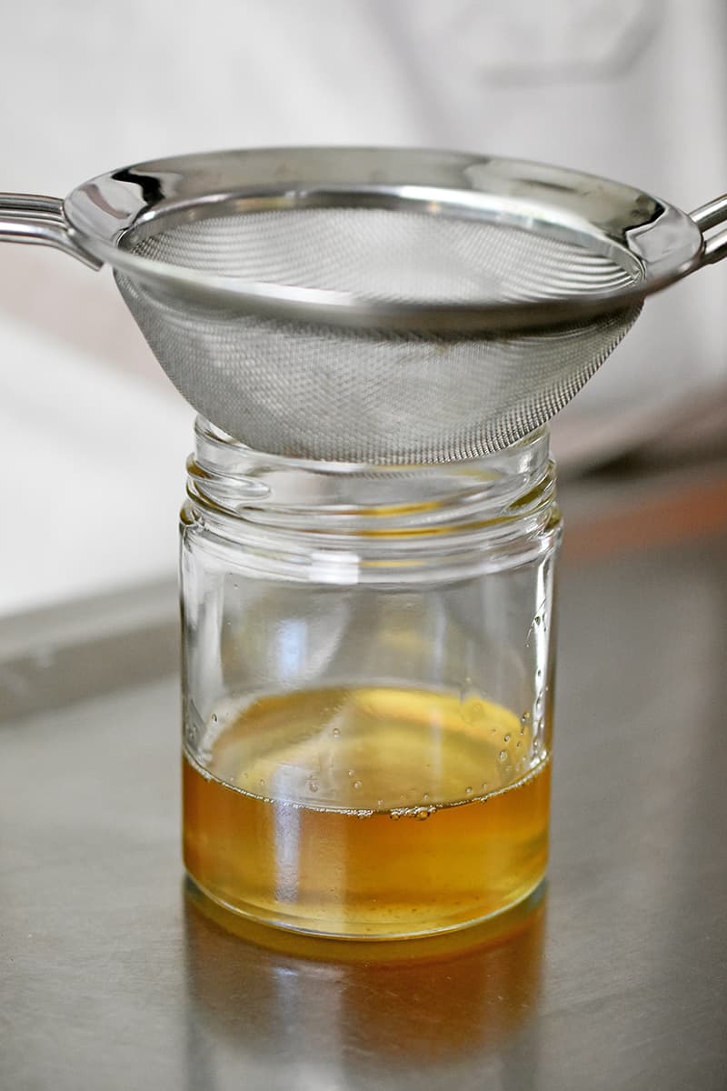 A side shot of a small glass jar filled with golden bacon drippings. The jar has a fine mesh sieve on top.