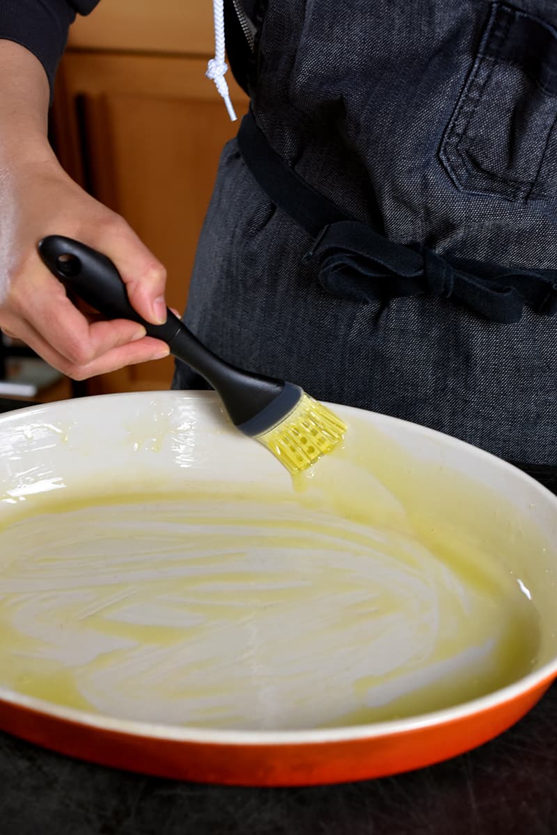 A hand is using a silicone brush to coat an enamel cast iron casserole pan with extra virgin olive oil