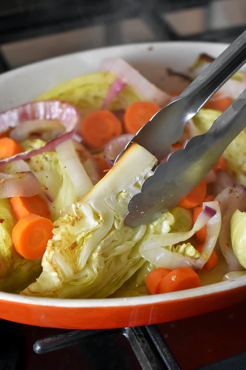 Metal tongs are turning over wedges of braised cabbage in a casserole dish