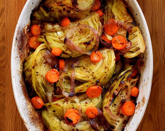 An overhead shot of the world's best braised green cabbage, carrot coins, and sliced onions in an oval casserole pan. Two hands are holding the handles with towels.