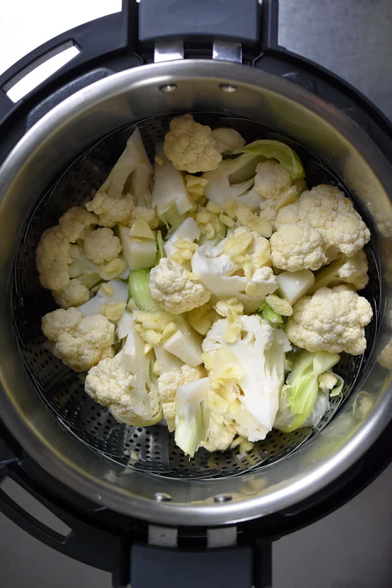 An overhead shot of cauliflower florets and sliced garlic in a steamer in an Instant Pot.