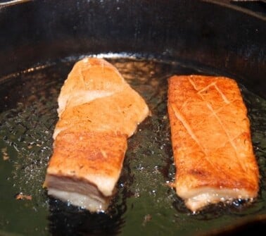Two sous vide cooked pork bellies frying on a cast iron skillet.