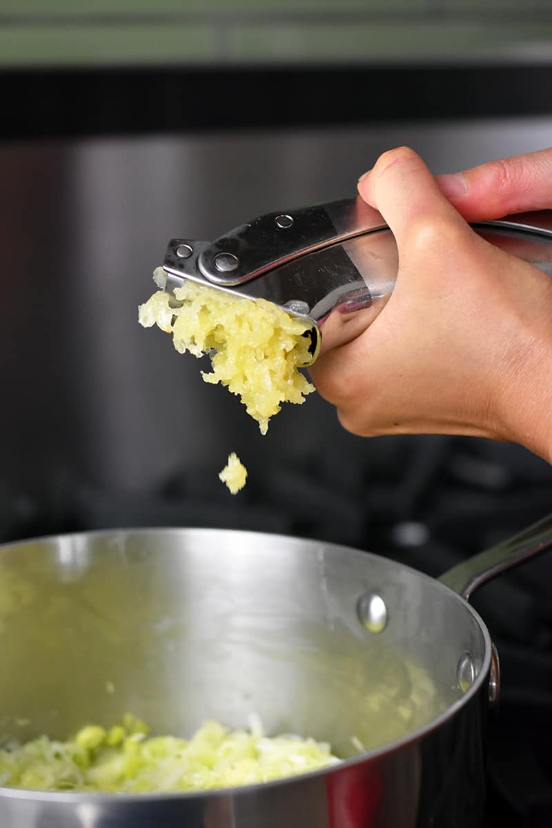 Adding minced garlic from a garlic press to a saucepan filled with softened leeks