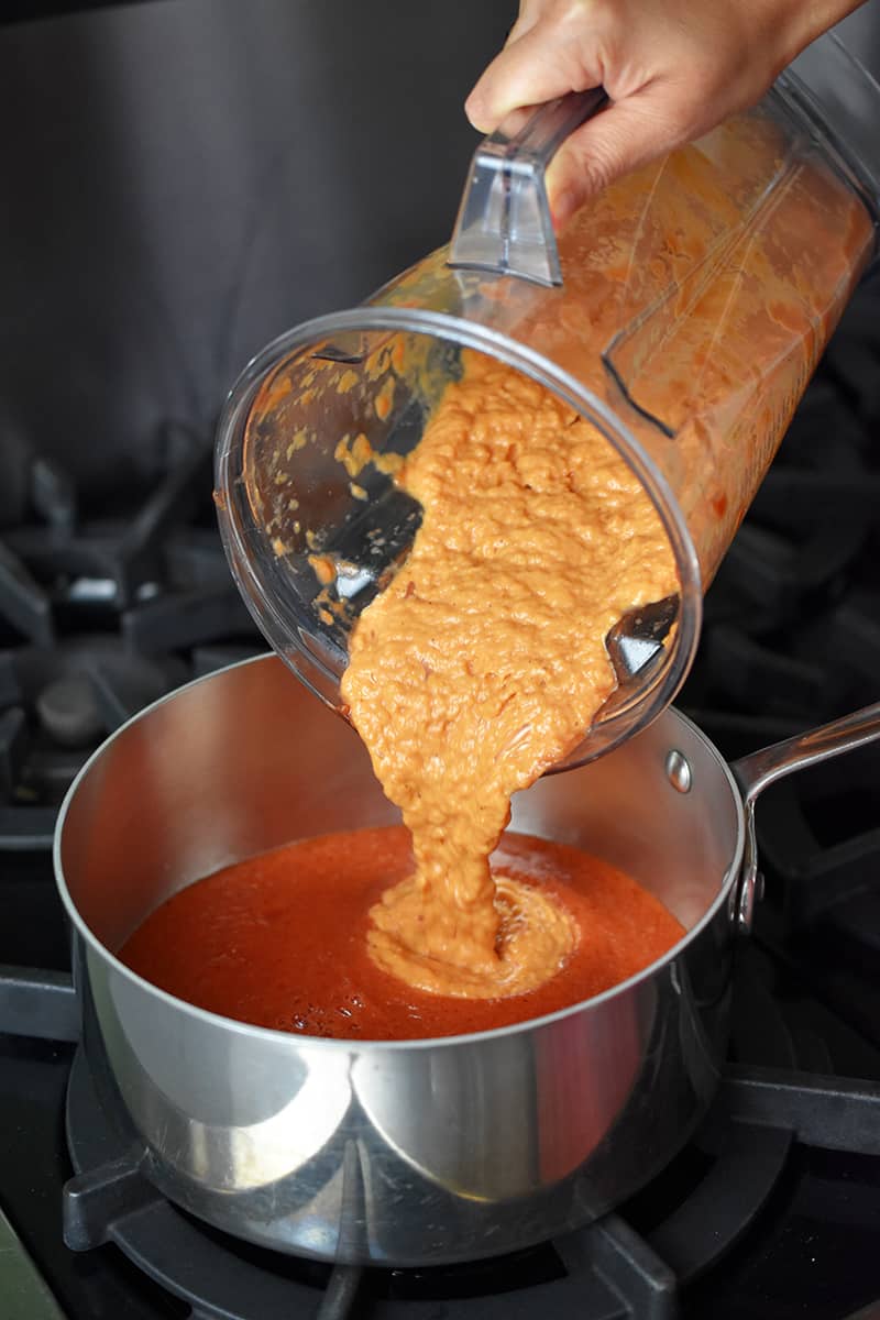 A blender filled with pureed leeks and tomatoes is being poured into a saucepan with pureed tomatoes.