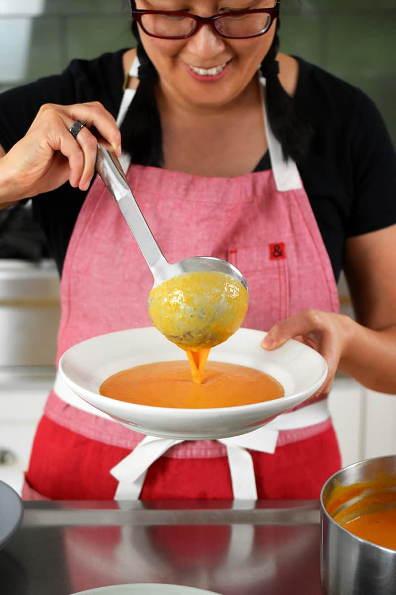 A smiling Asian woman is ladling cream of tomato soup into a bowl.