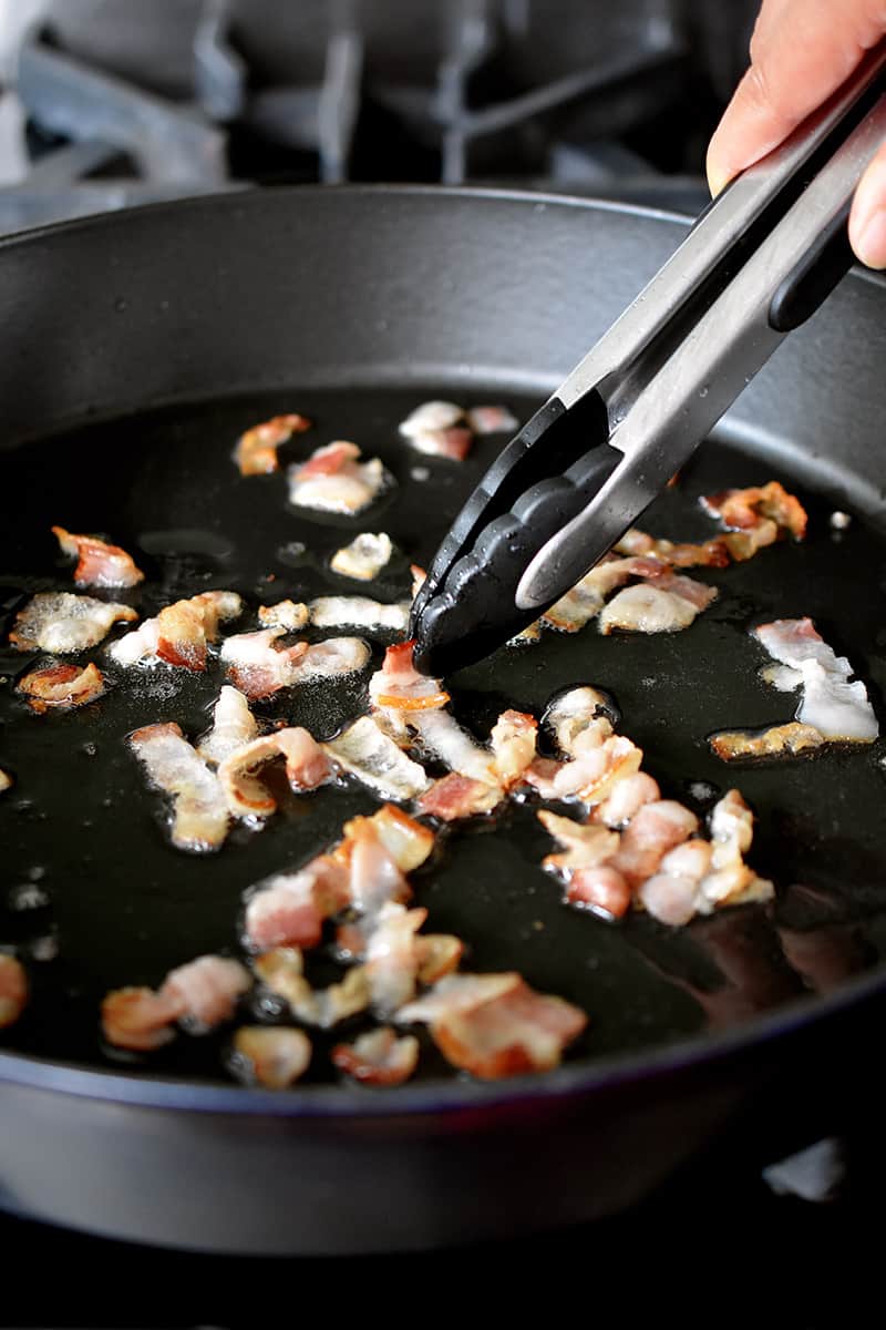 A pair of tongs is pushing raw bacon bits in a large cast iron skillet.