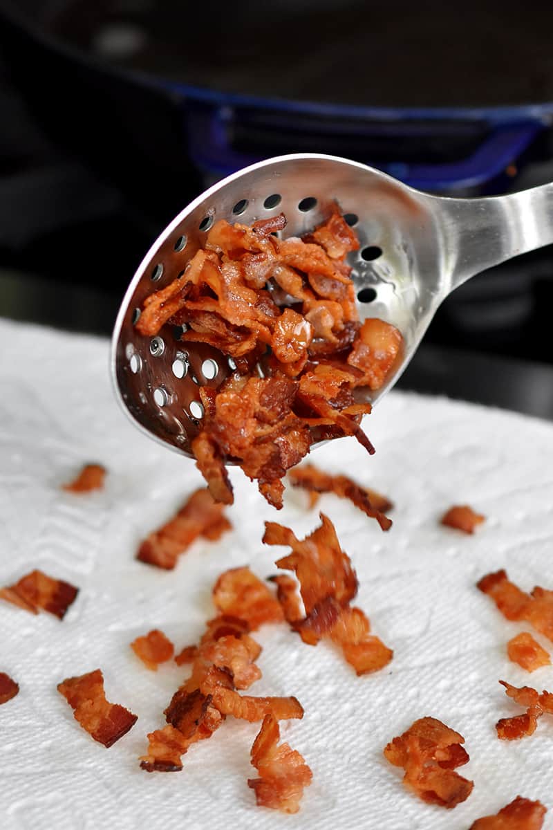 A metal slotted spoon is transferring crispy bacon bits on a paper towel.