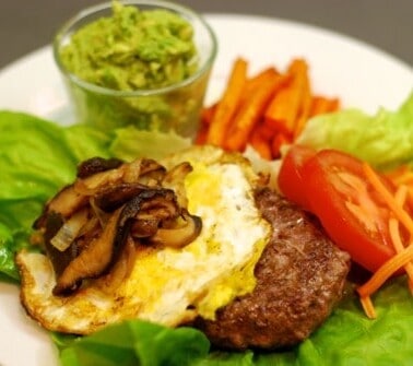 A plate with a lamb burger on top of a lettuce "bun" topped with a fried egg and sautéed shiitake mushrooms and onions.