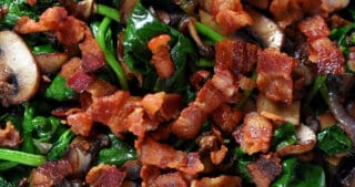 An overhead shot of a plate filled with sautéed spinach, crispy bacon bits, shallots, and mushrooms.