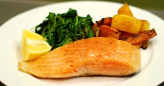 A plate of sous vide wild king salmon with a lemon wedge, sautéed spinach, and carrots.