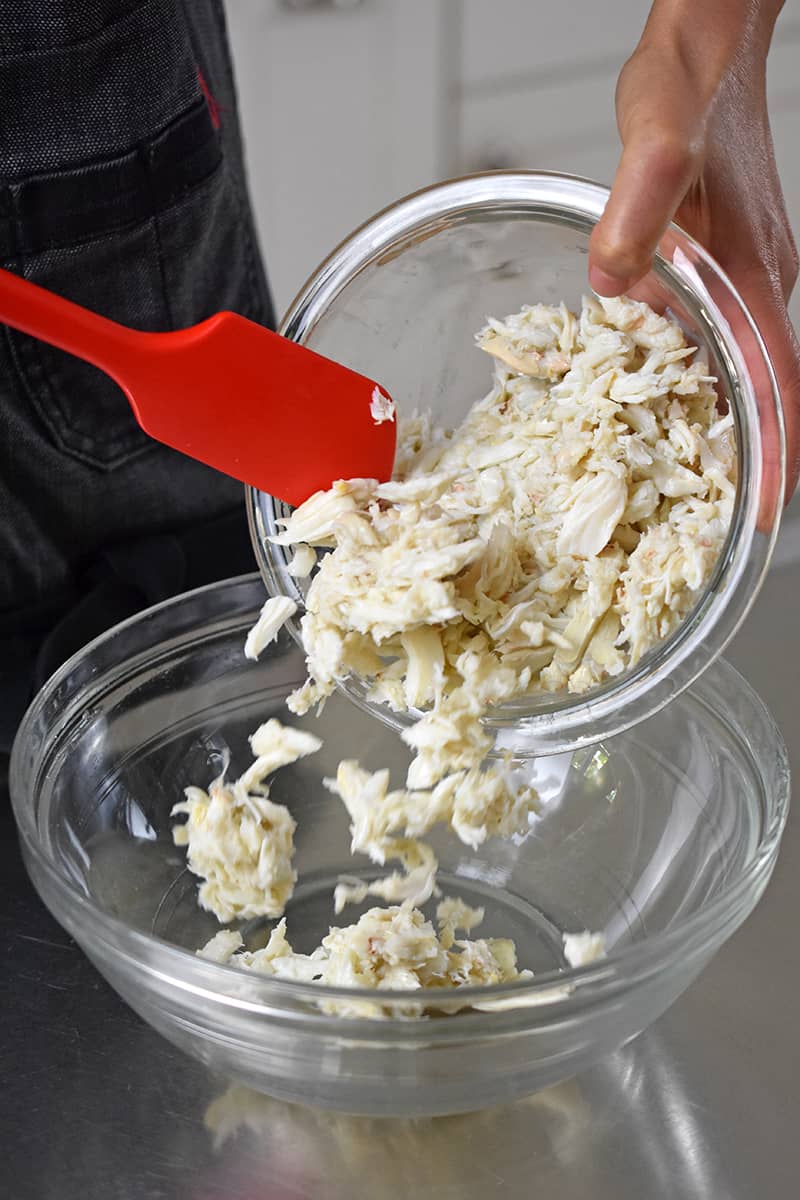 A red spatula is transferring fresh crab meat to a large glass bowl.