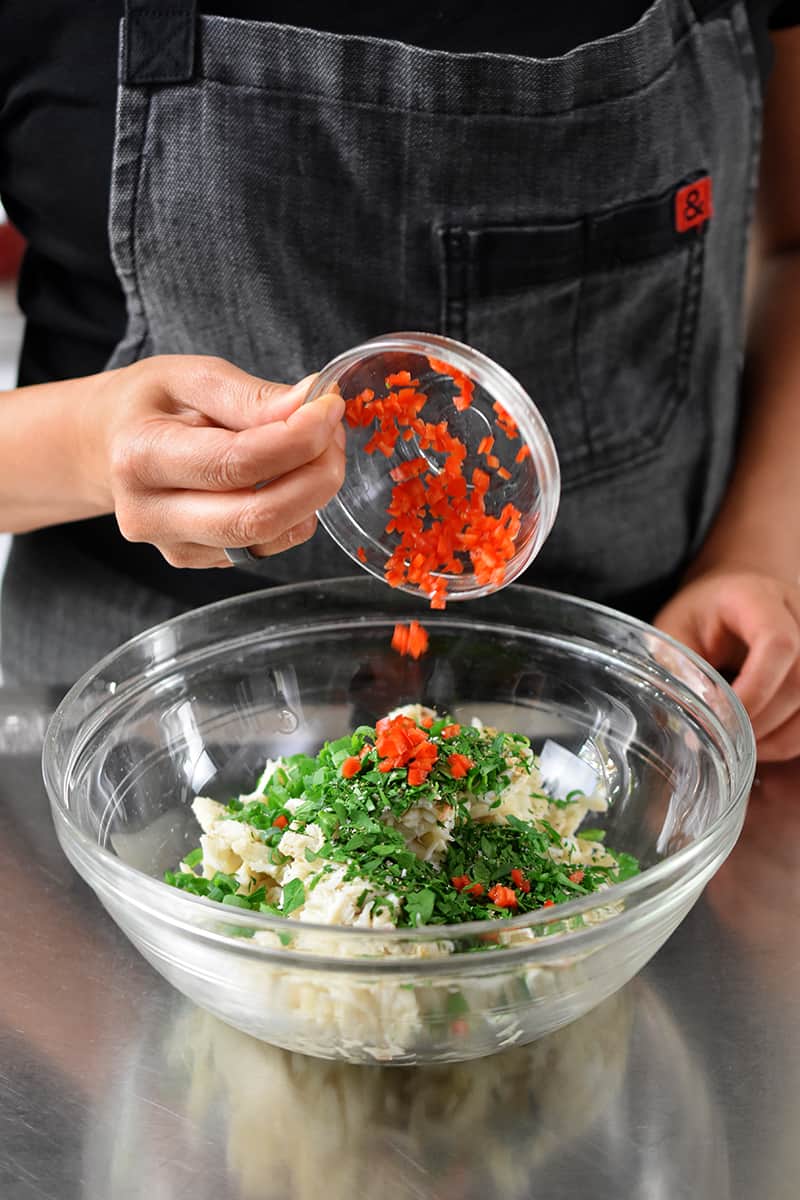 Someone in a gray apron is adding diced red peppers to a bowl filled with crab salad ingredients.