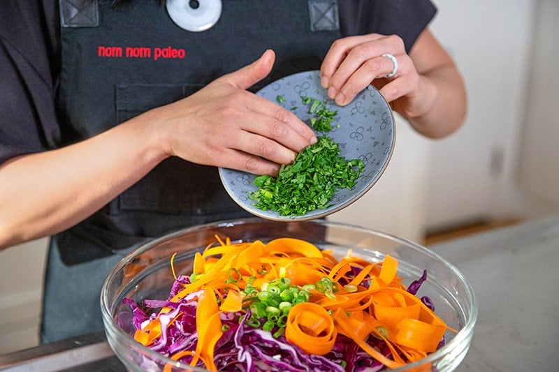 A shot of someone adding cilantro and green onions to a bowl filled with red cabbage and carrots to make red cabbage slaw.