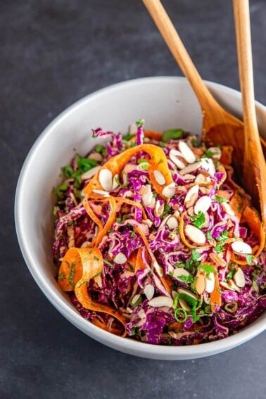 A shot of Whole30 and paleo red cabbage slaw in a large white bowl with two wooden serving spoons inside.