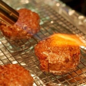 Hamburger patties made out of lamb are sitting on a wire rack. One patty has a kitchen blow torch blowing fire on it.