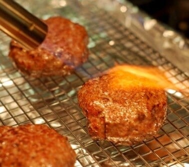Hamburger patties made out of lamb are sitting on a wire rack. One patty has a kitchen blow torch blowing fire on it.
