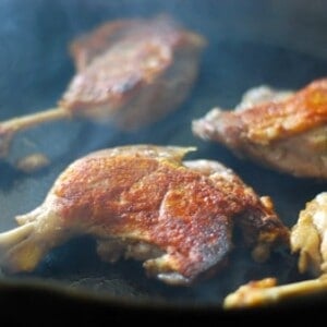 Four duck legs cooking on a cast iron skillet with smoke rising off the ducks.