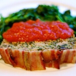 A spinach and mushroom meatloaf topped with bacon is on a plate with cooked spinach and it is topped with marinara sauce.