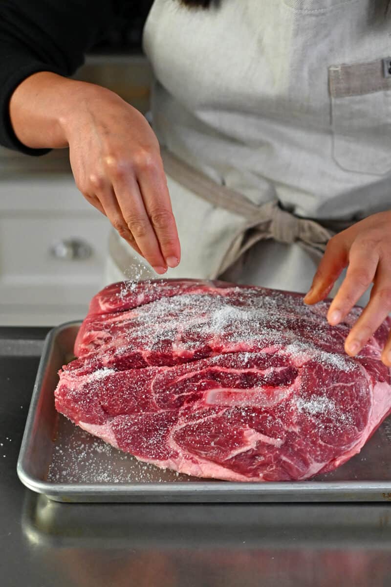 A hand is sprinkling kosher salt all over a large bone-in pork shoulder roast.