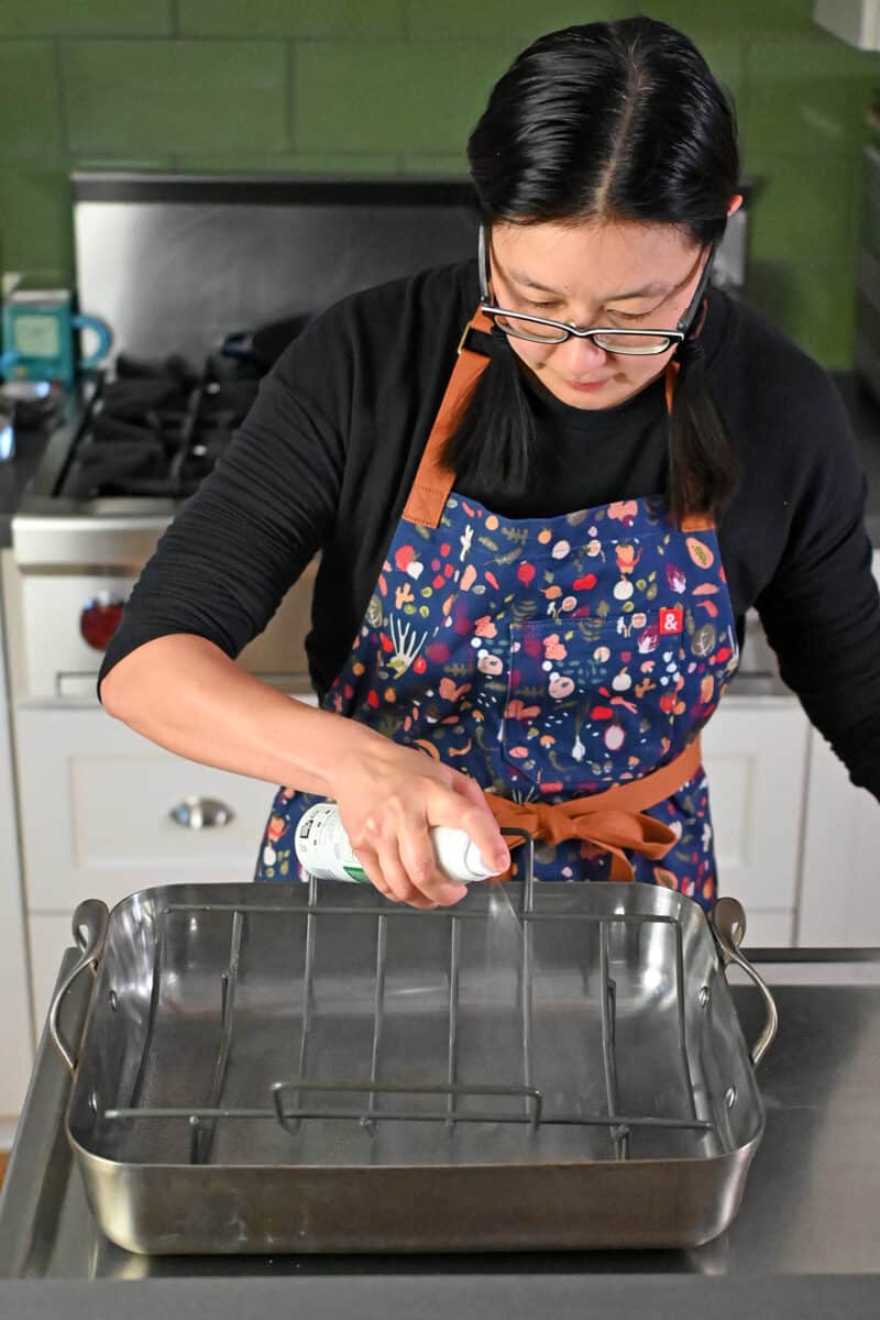 An Asian woman in glasses is spraying avocado oil on a rack in a roasting pan.