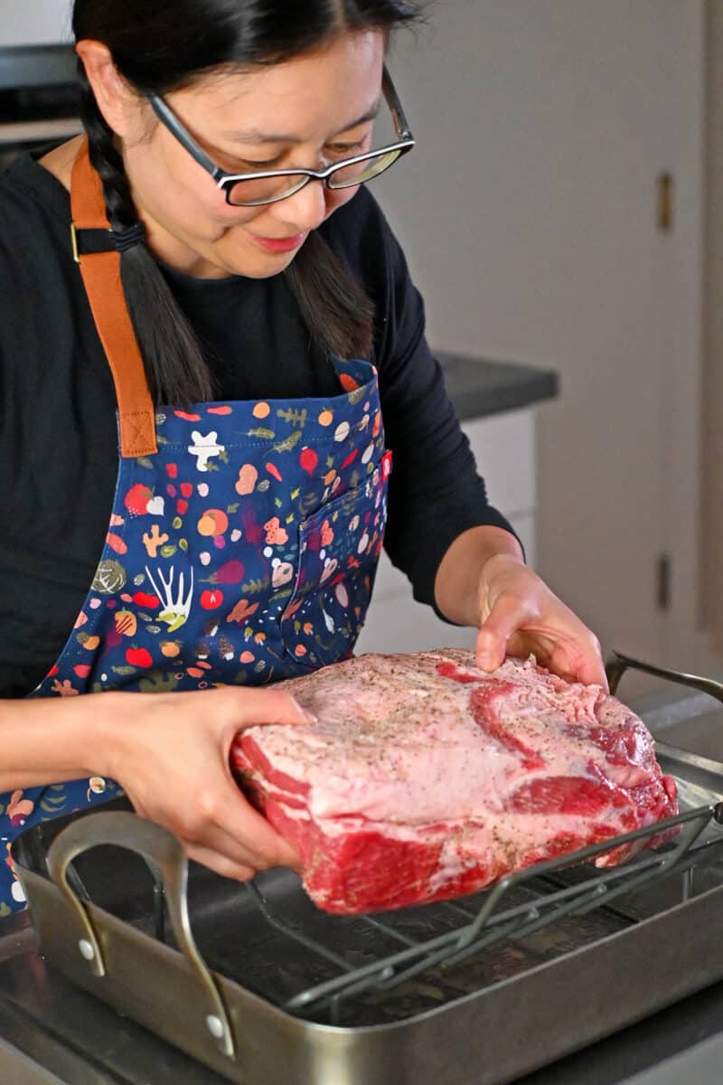 An Asian woman is placing a seasoned bone-in pork shoulder roast onto a greased rack in a roasting pan. 