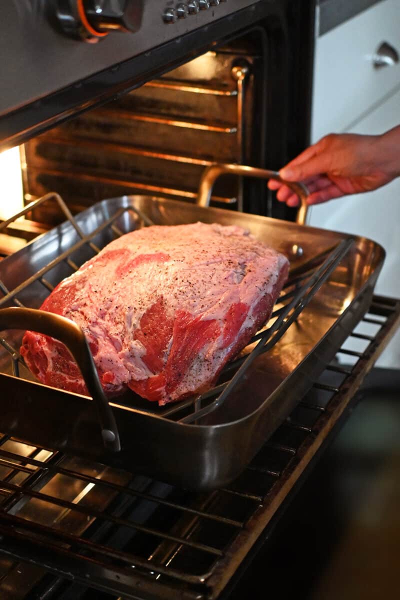 Placing a roasting pan with a seasoned bone-in pork shoulder roast into an open oven.