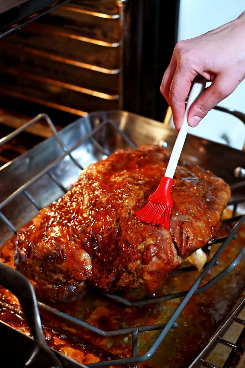 A silicone brush is adding a peach jam glaze onto a golden brown pork shoulder roast in the oven.