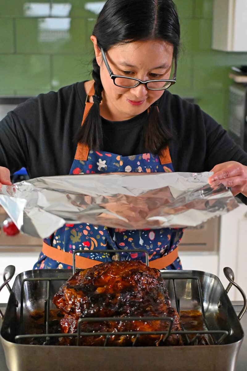 An Asian woman in glasses is placing a large piece of aluminum foil on top of the slow roasted pork shoulder. 