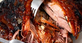 Two forks are shredding a pork shoulder roast that is topped with a peach barbecue sauce.