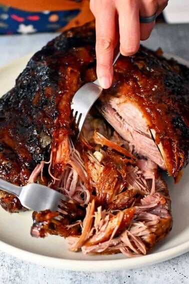 Two forks are shredding a pork shoulder roast that is topped with a peach barbecue sauce.