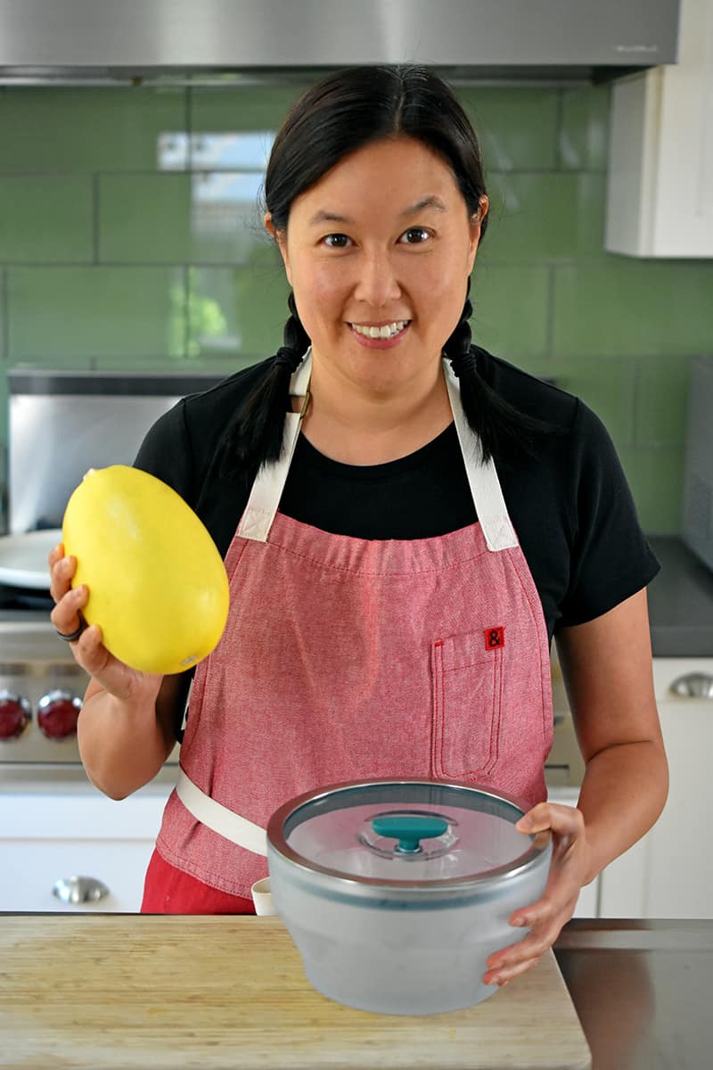 A smiling Asian woman is holding a small spaghetti squash and a large deep Anyday microwave bowl.