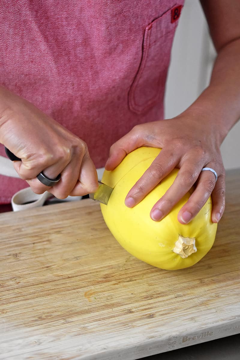 A person in a red apron is using a sharp paring knife to cut a small spaghetti squash in half crosswise in the middle. 