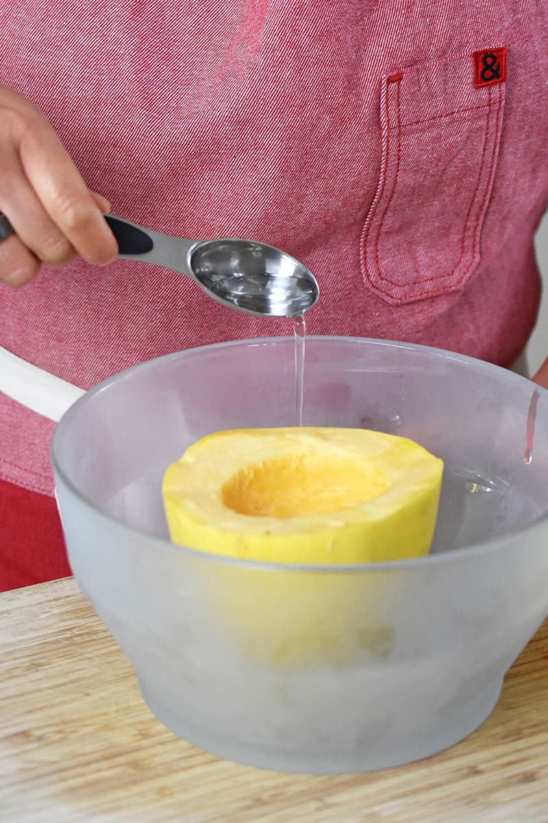 Someone in a red apron is adding a spoonful of water to a glass container with a half of a spaghetti squash inside