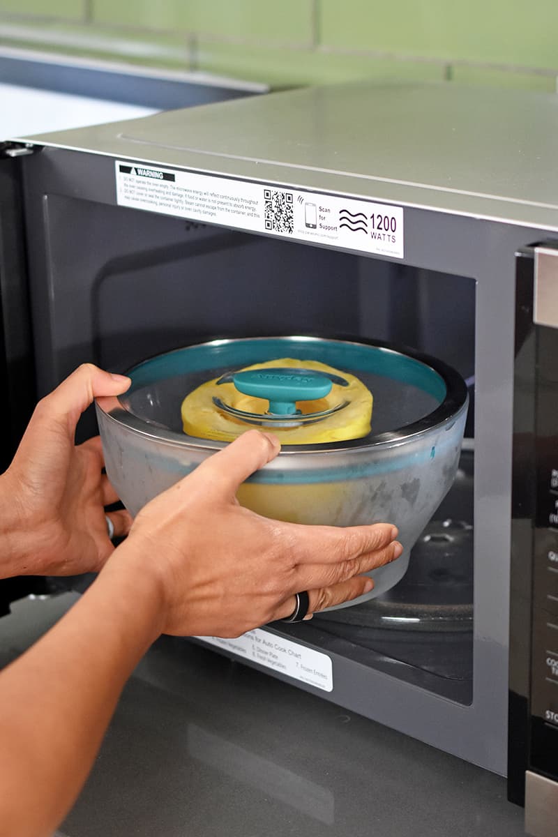 Two hands are placing a glass bowl filled with a halved spaghetti squash into an open microwave