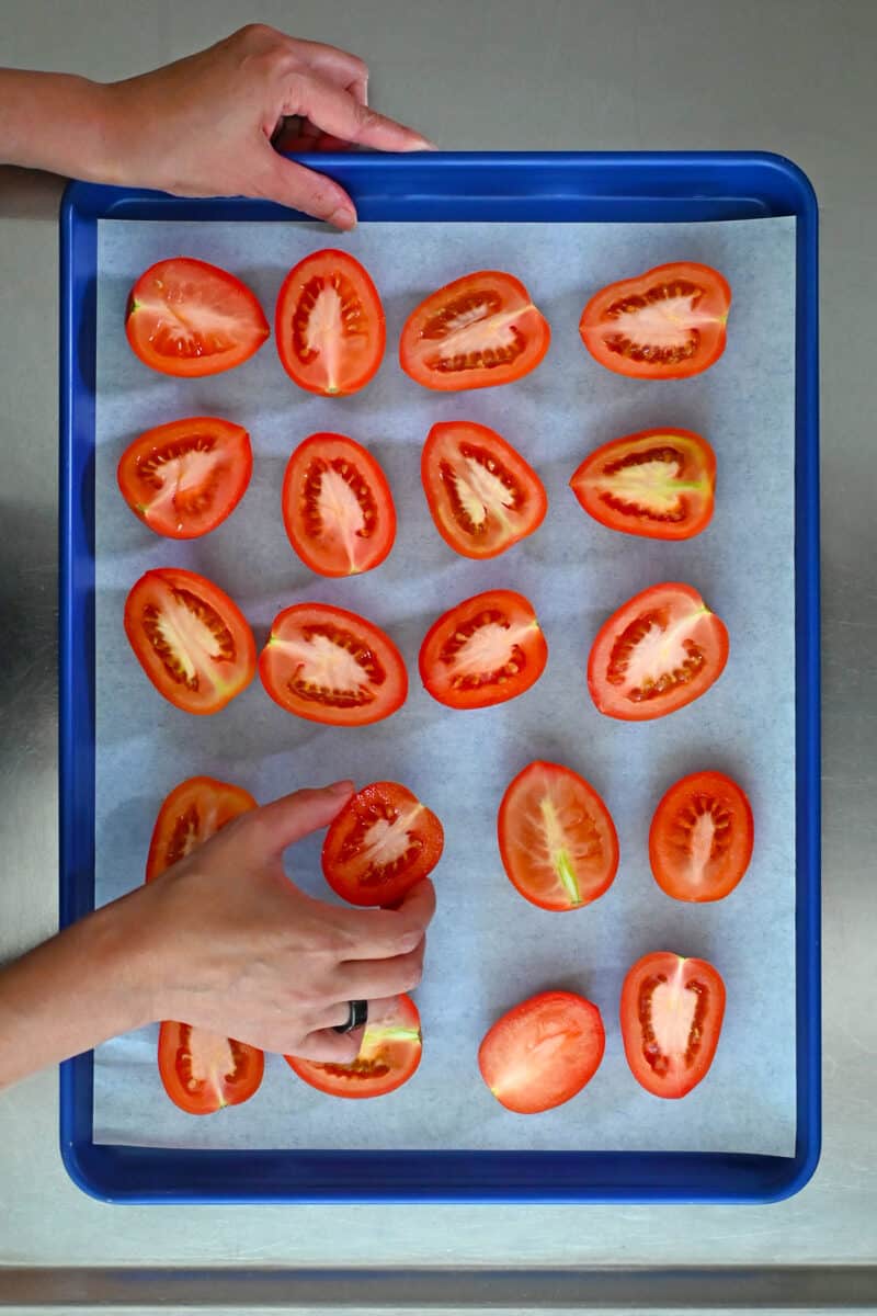 An overhead shot of two hands arranging halved Roma tomatoes on a blue baking sheet.