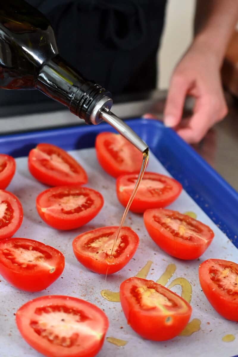 Drizzling extra virgin olive oil on halved tomatoes on a blue baking sheet.