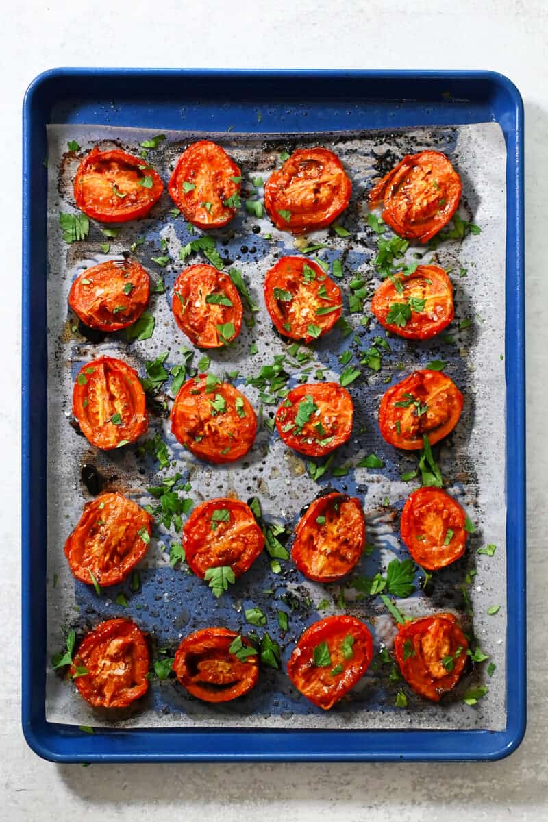 An overhead shot of a blue baking sheet with oven roasted tomatoes drizzled with balsamic vinegar and fresh herbs.