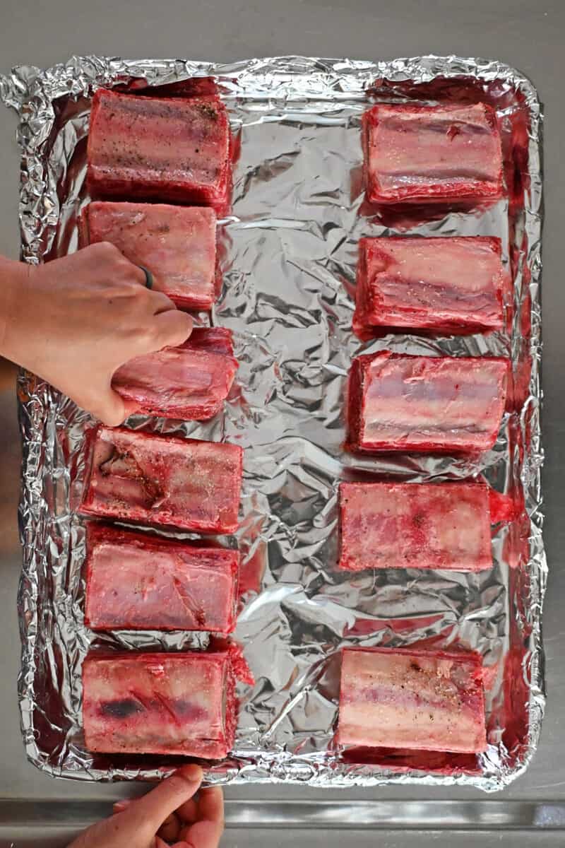 A hand is placing seasoned raw beef short ribs, bone-side up, on a foil lined rimmed baking sheet.