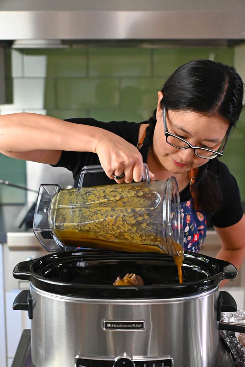 An Asian woman in glasses is pouring a brown sauce into an open slow cooker.