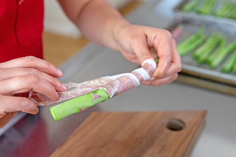 A person is wrapping a thin strip of prosciutto around an asparagus spear.