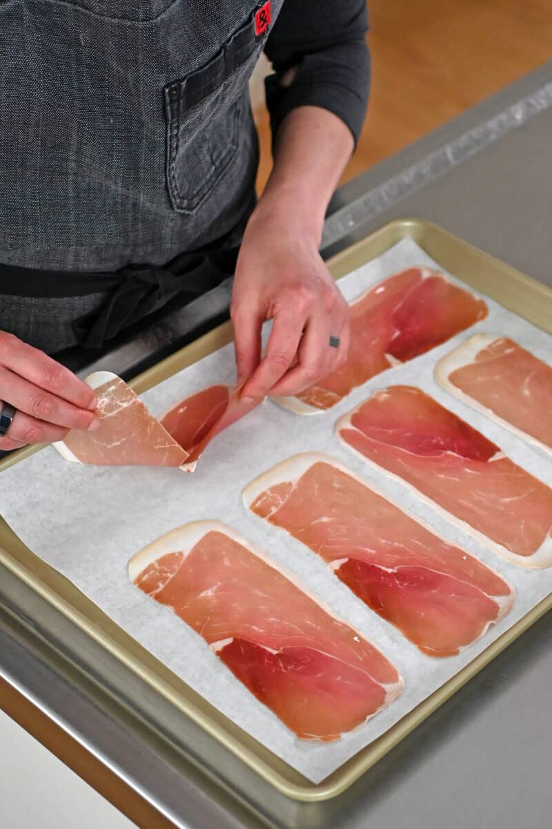 Two hands are placing thinly sliced prosciutto slices on a parchment-lined rimmed baking sheet to make crispy prosciutto
