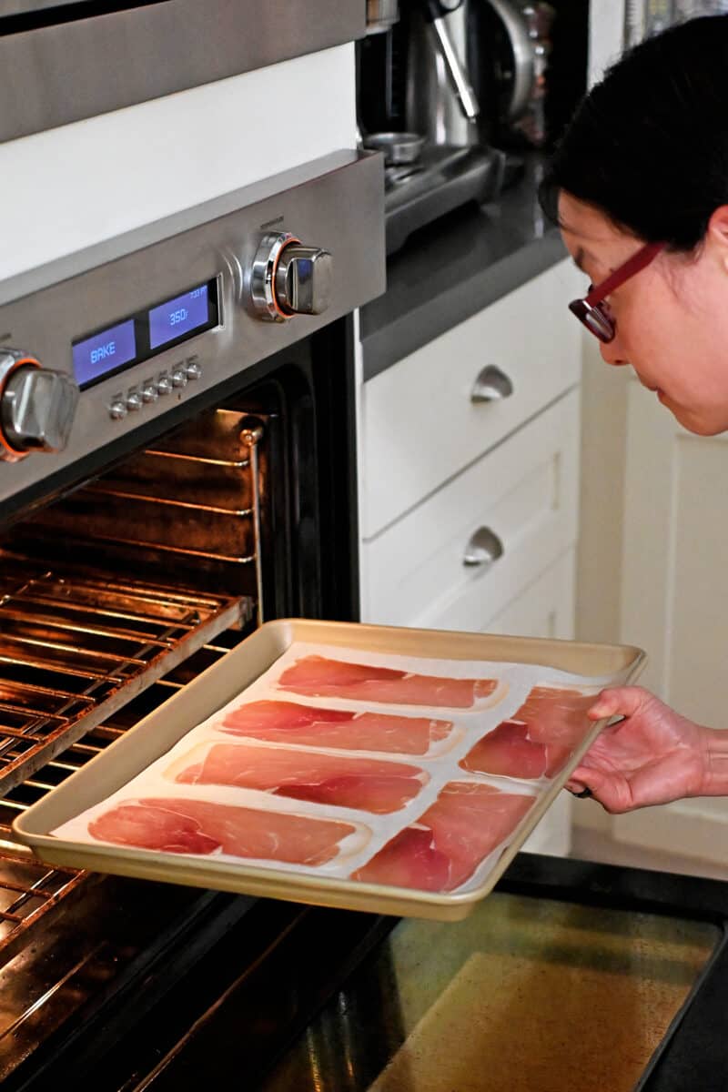 An Asian woman in glasses is placing a rimmed baking sheet with prosciutto slices into the oven,