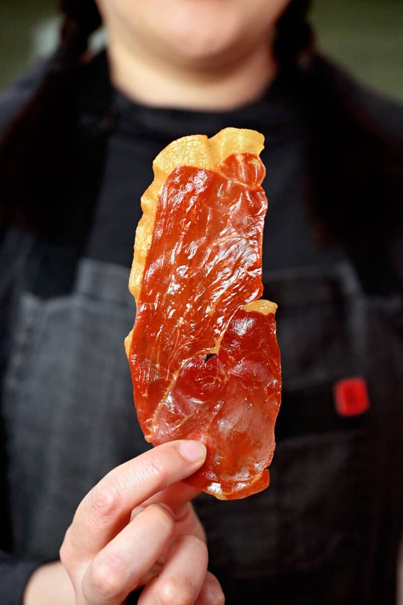 A person in a gray apron is holding up a crispy prosciutto chip