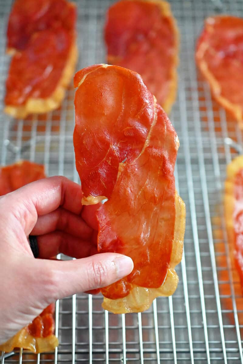 A hand is holding a crispy prosciutto chip that was picked up from a metal cooling rack