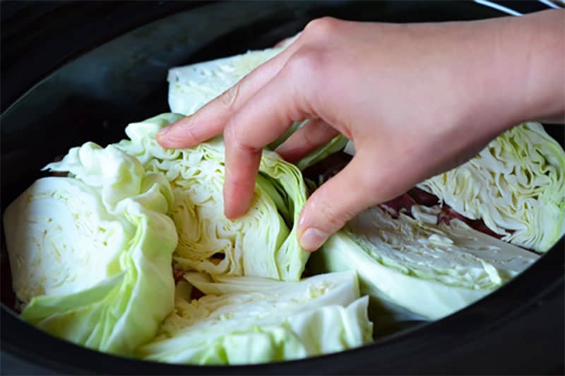 A hand is placing cabbage wedges on top of Slow Cooker Cheater Pork Stew in an open Crock-Pot