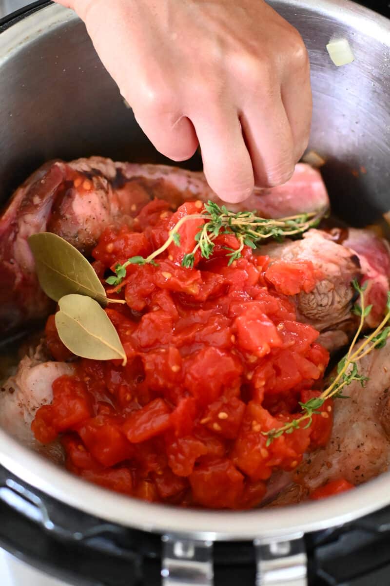 Adding dried bay leaves and fresh herbs to an Instant Pot filled with lamb shanks.