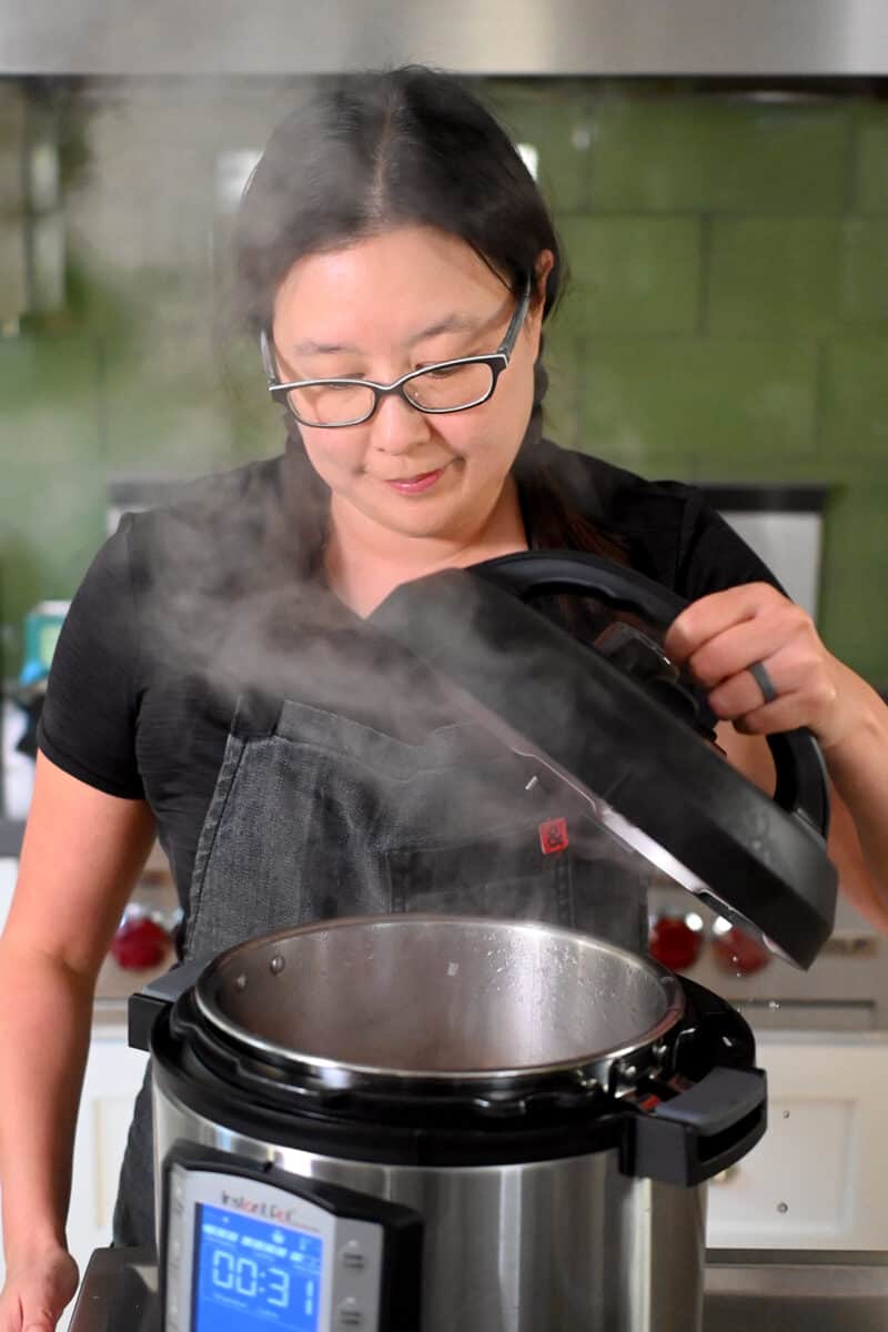 An Asian woman in glasses is opening an Instant Pot and lots of hot steam is coming out.