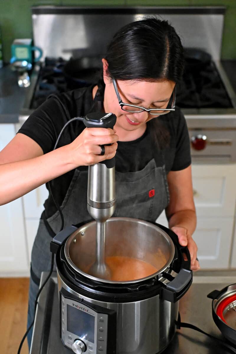 A smiling Asian woman in glasses is using an immersion blender to blend a sauce in an Instant Pot.