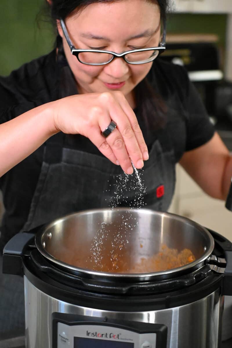 An Asian woman in glasses is seasoning the sauce for lamb shanks in an Instant Pot.