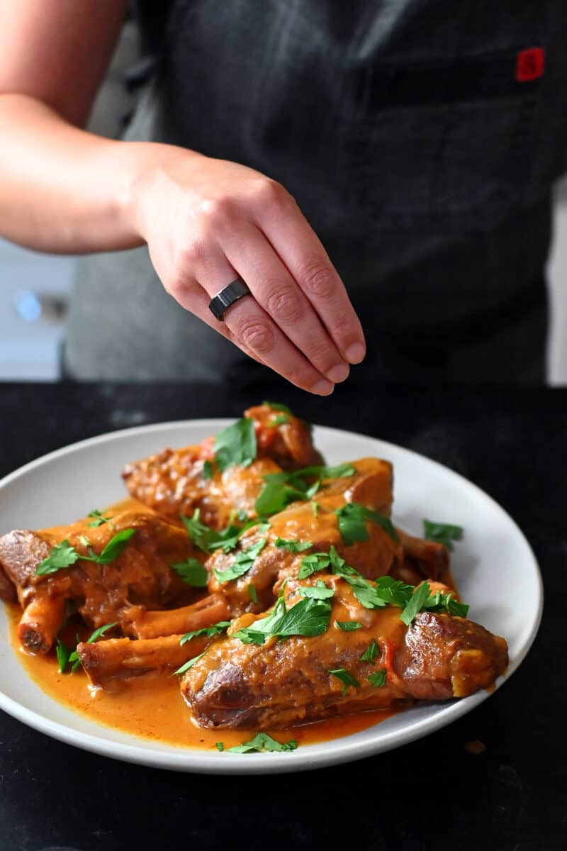 A hand is sprinkling fresh Italian parsley on some lamb shanks cooked in an Instant Pot.